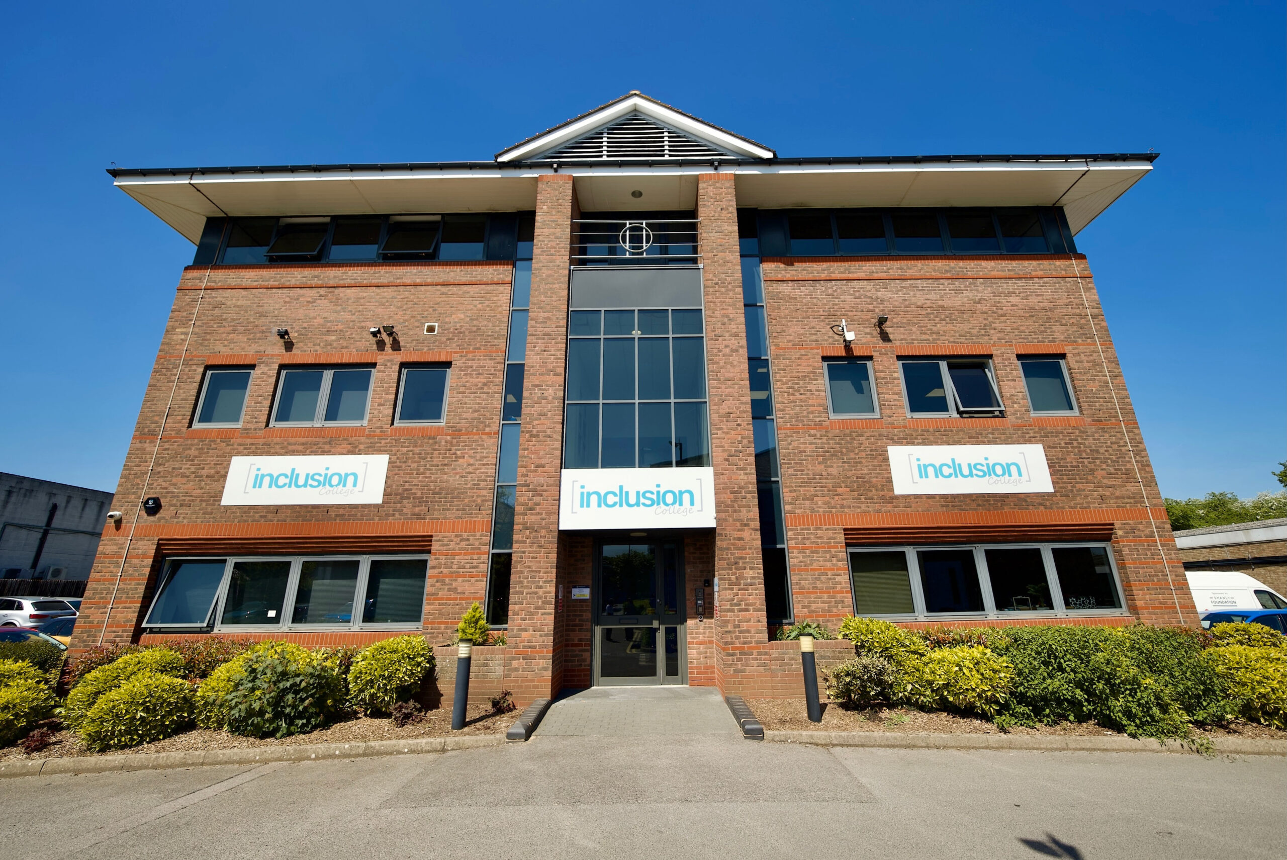 Brick campus building with a glass entrance and signs reading 'inclusion college' against a blue sky behind hedge