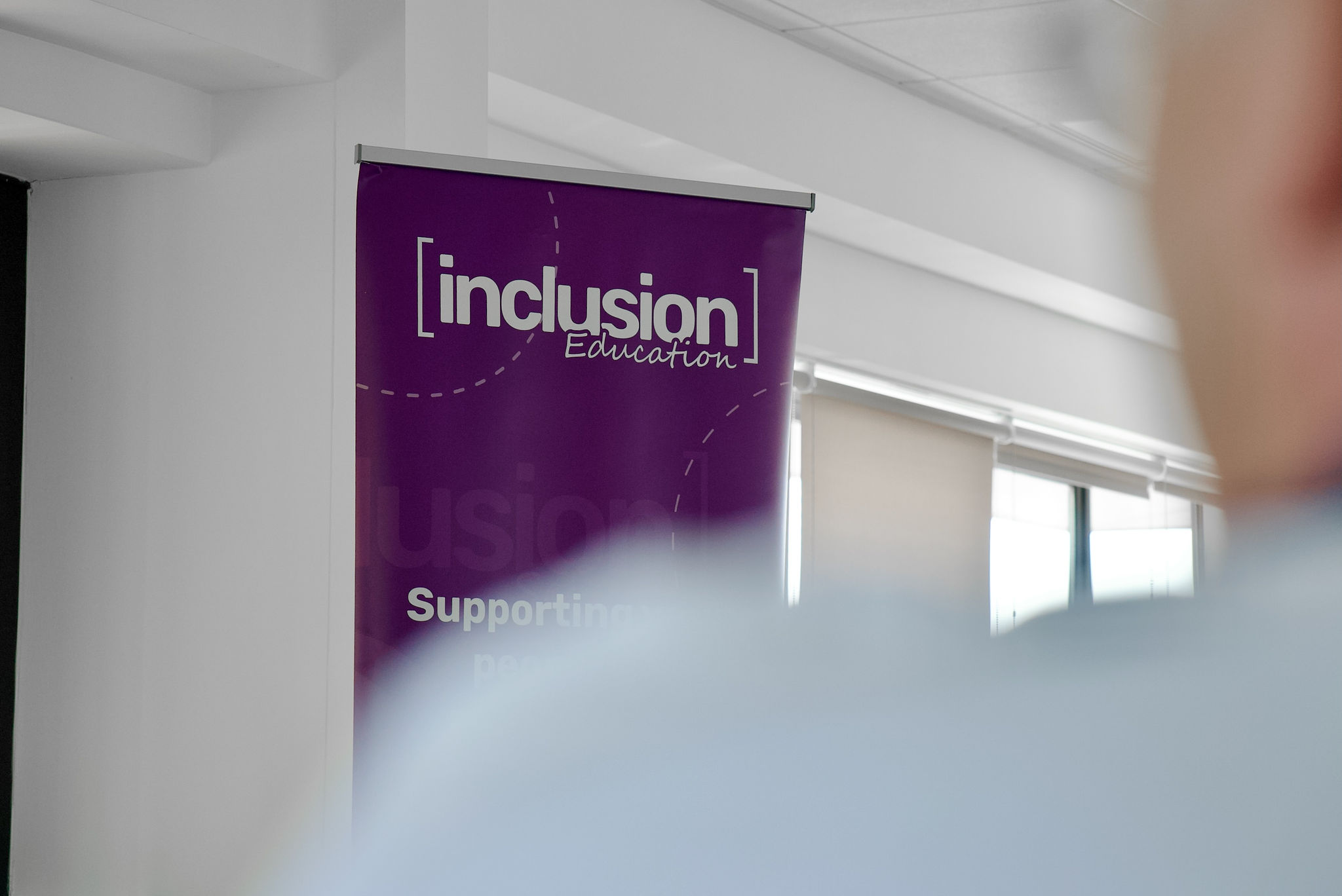 Purple promotional banner reading 'Inclusion Education' hanging in a bright room with white walls and windows, partially obscured by frosted foreground.