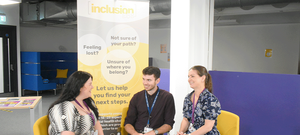 Three people sit in yellow chairs having a discussion in front of an 'inclusion' banner.